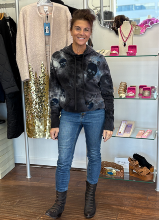 Woman in a store wearing a tie-dye cashmere hoodie with  blue jeans, and brown boots.