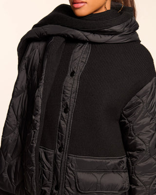 Person wearing a black quilted jacket and scarf on a beige background